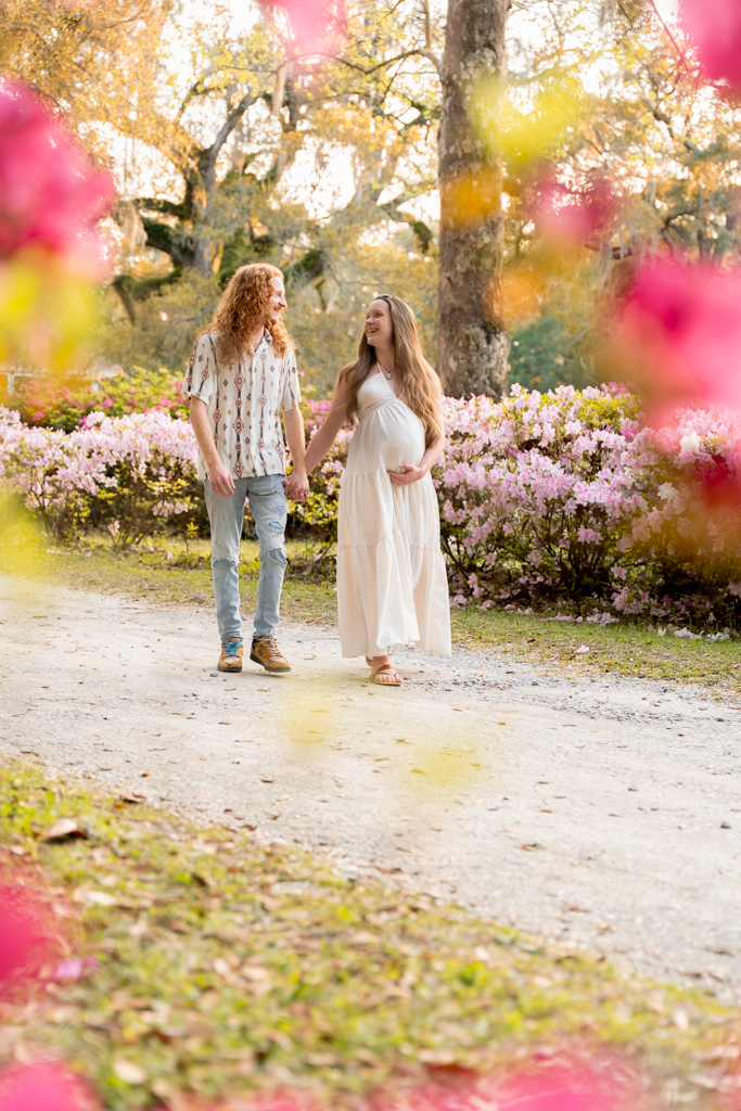 Expecting couple walking hand-in-hand along a pathway lined with azaleas and oak trees at Eden Gardens.
